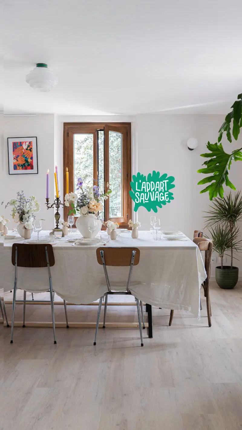 La salle de L’Appart Sauvage : une longue table nappée de blanc, bougies et bouquets, chaises de bistrot, grande fenêtre et plantes vertes.