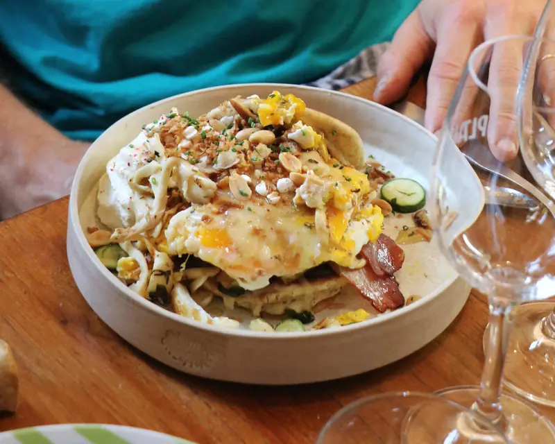 Une assiette creuse de brunch : œuf au plat sur un pancake, lard grillé, concombre, fromage frais, cacahuètes et oignons frits, à côté d’un verre à vin.