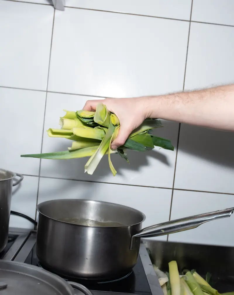 Une main jette des poireaux dans une casserole fumante, devant un mur carrelé de blanc.