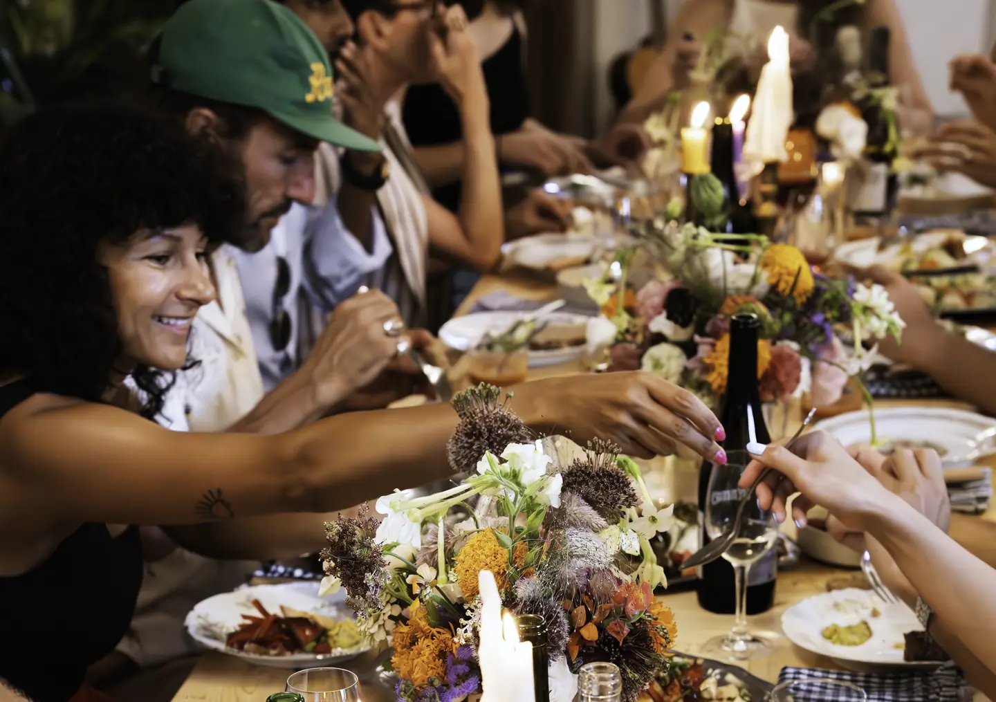 Une grande tablée à L’Appart Sauvage : des amis rient autour d’une longue table de bois, bougies, bouquets de fleurs des champs, bouteilles et plats à partager.